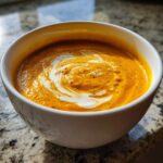 A close-up of rich orange squash soup topped with a swirl of cream and cracked black pepper.