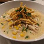 A close-up of creamy tortilla soup in a white bowl, featuring shredded chicken, corn, and crispy tortilla strips.