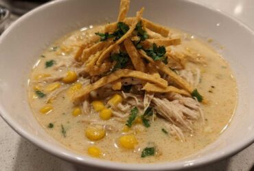 A close-up of creamy tortilla soup in a white bowl, featuring shredded chicken, corn, and crispy tortilla strips.