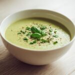A close-up of a bowl of creamy zucchini soup, garnished with fresh basil leaves and cracked pepper.