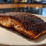 A close-up of a thick fillet of blackened salmon with a dark, spicy crust resting on a white plate.