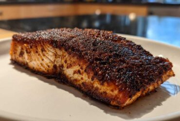 A close-up of a thick fillet of blackened salmon with a dark, spicy crust resting on a white plate.