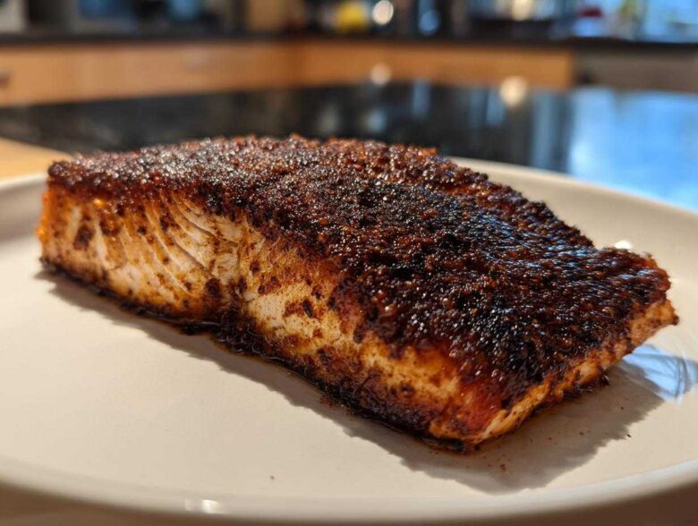 A close-up of a thick fillet of blackened salmon with a dark, spicy crust resting on a white plate.