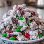 A mound of powdered sugar-coated christmas puppy chow mixed with red and green candies on a white plate.