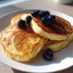 Stack of three golden brown coconut flour pancakes topped with fresh blueberries and syrup on a white plate.