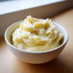 A close-up of fluffy mashed potatoes in a white bowl, seasoned generously with cracked black pepper.