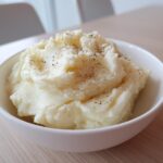 A close-up of a generous serving of fluffy mashed potatoes topped with cracked black pepper in a white bowl.