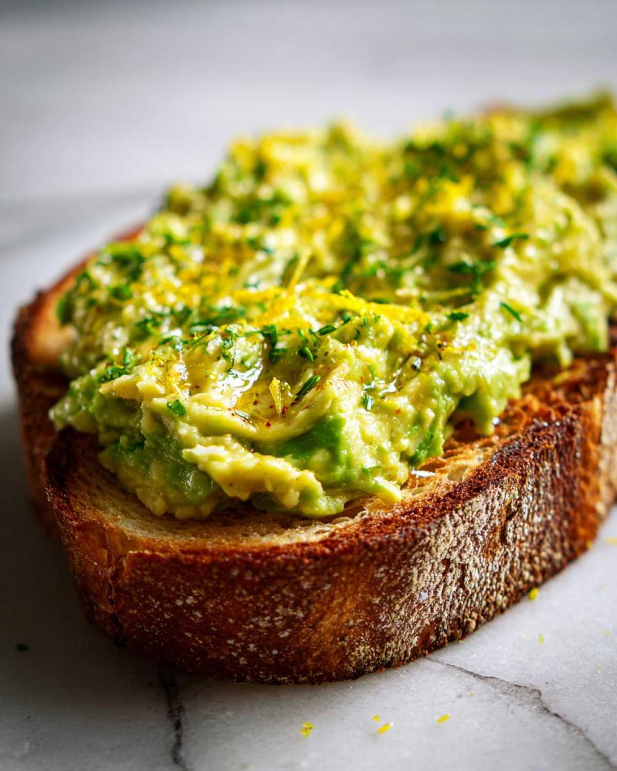 Close-up of a thick slice of toasted bread topped with mashed avocado toast, lemon zest, and herbs.