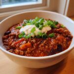 A close-up of a hearty bowl of rich pumpkin chili topped with sour cream and chopped green onions.