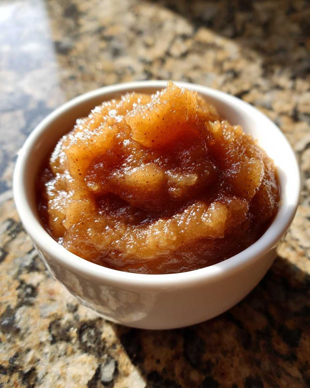 Close-up of rich, spiced homemade applesauce served in a small white bowl on a granite countertop.