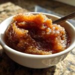 A small white bowl filled with richly colored, spiced homemade applesauce, served with a spoon.