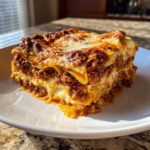 A thick, cheesy slice of homemade lasagna featuring rich meat sauce and pasta layers, served on a white plate.