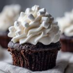 A close-up of a rich, dark chocolate hot chocolate cupcake topped with white frosting and mini marshmallows.