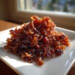 A mound of deeply browned, glossy caramelized onions piled on a square white plate, catching the sunlight.