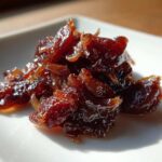 Close-up of a small pile of deep brown, glossy caramelized onions on a white square plate.