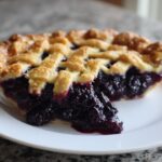 A close-up of a slice of juicy blackberry pie featuring a golden lattice crust and rich, dark berry filling.