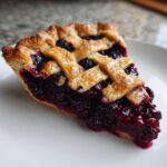 A close-up of a juicy slice of blackberry pie featuring a golden lattice crust and deep purple filling.