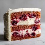 Close-up of a moist slice of cherry cake with two layers of dark cake, white cream filling, and whole cherries.