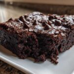 A close-up of a rich, fudgy slice of black bean brownies topped with melted chocolate chips on a white plate.