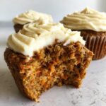 Close-up of a bitten carrot cake muffin showing moist texture and shredded carrots, topped with thick cream cheese frosting.