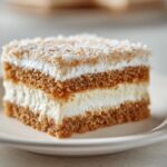 Close-up of a layered slice of moist coconut cake featuring brown cake layers, white cream filling, and toasted coconut topping.