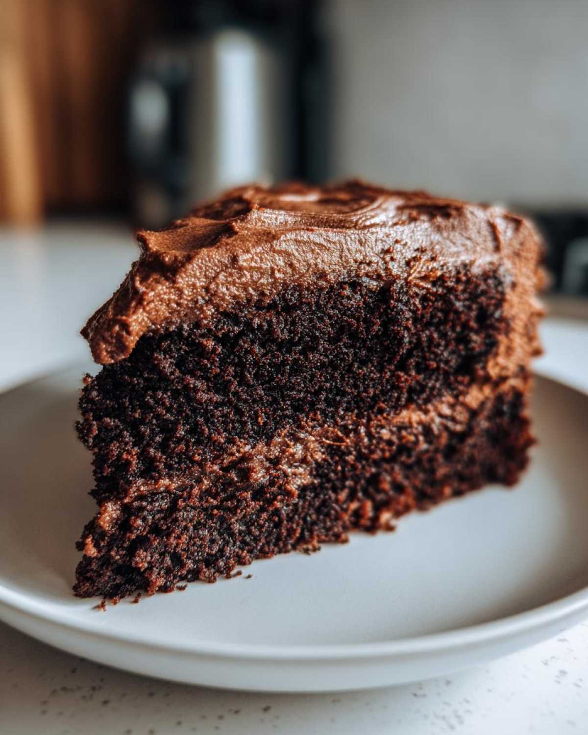 Close-up of a moist slice of homemade chocolate cake topped with thick, dark chocolate frosting on a white plate.