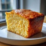 A close-up of a thick, moist slice of golden sourdough cornbread on a white plate.