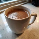 Close-up of a white mug filled with rich, foamy crockpot hot chocolate sitting on a light countertop.
