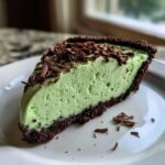 A close-up of a slice of vibrant green, creamy grasshopper pie with a dark chocolate crust, topped with chocolate shavings.