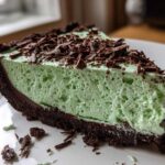 A close-up of a slice of bright green, fluffy grasshopper pie with a dark chocolate crust and topped with chocolate shavings.