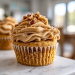 A close-up of a single, perfectly frosted pecan pie cupcake topped with chopped pecans and cinnamon.
