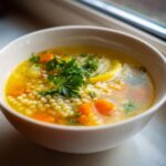 Close-up of a white bowl filled with bright yellow penicillin soup, featuring Israeli couscous, chopped carrots, and fresh parsley garnish.