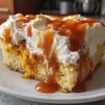 A close-up of a slice of moist pumpkin poke cake topped with whipped cream and drizzled heavily with caramel sauce.