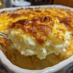 A spoonful of creamy, cheesy baked mashed potatoes being lifted from a casserole dish with a golden brown crust.