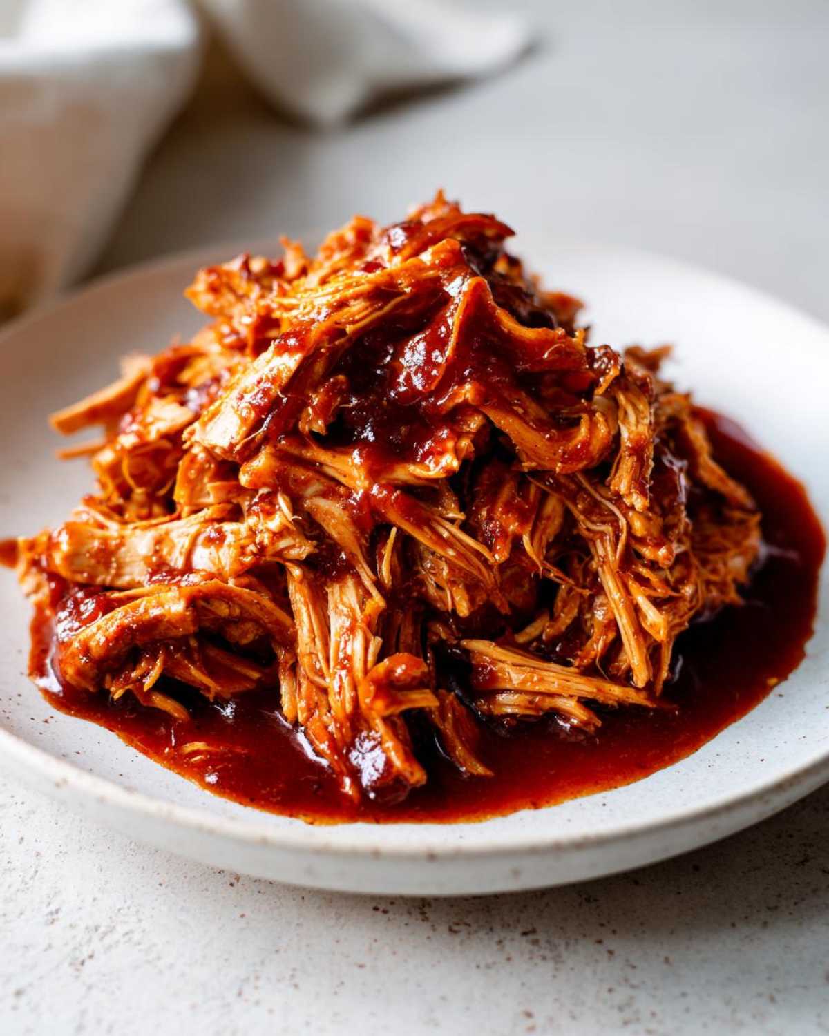 Close-up of shredded cranberry chicken coated in a thick, dark red sauce, served on a light plate.
