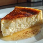 A close-up of a thick slice of silky egg custard with a caramelized top, resting in syrup on a white plate.