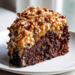 A close-up of a moist slice of german chocolate poke cake topped with rich coconut-pecan frosting.
