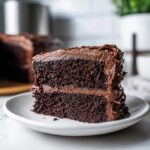A close-up of a rich, dark slice of homemade chocolate cake layered with creamy chocolate frosting.