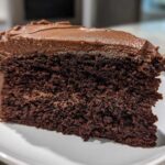 Close-up of a rich, dark slice of homemade chocolate cake topped with thick chocolate frosting on a white plate.
