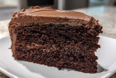 Close-up of a rich, dark slice of homemade chocolate cake topped with thick chocolate frosting on a white plate.