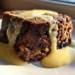 Close-up of a rich, dark slice of traditional plum pudding generously topped with flowing yellow custard sauce.