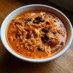 Close-up of a white bowl filled with creamy, orange-red slow cooker buffalo chicken chili topped with dark kidney beans.