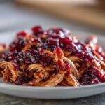 Close-up of shredded slow cooker cranberry chicken topped with glossy, dark red cranberry sauce on a white plate.