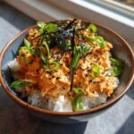 Close-up of a vibrant tuna rice bowl topped with spicy orange tuna mixture, green onions, nori, and black sesame seeds.