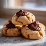 A stack of three golden, sugar-coated peanut butter blossoms topped with a swirl of rich, dark chocolate frosting.