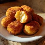 A stack of golden brown, crispy potato croquettes served on a white plate, one is broken open showing the soft interior.