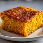 A square slice of vibrant orange sweet potato cobbler with a caramelized, dark brown top, served on a white plate.