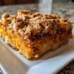 A close-up of a square slice of sweet potato cobbler featuring a bright orange filling and thick, brown sugar crumb topping.