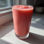 A tall glass filled with a frosty, bright pink watermelon smoothie sitting on a white counter near a window.