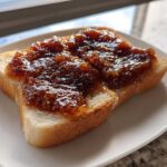 A thick slice of white toast generously topped with dark, glistening homemade apple butter.